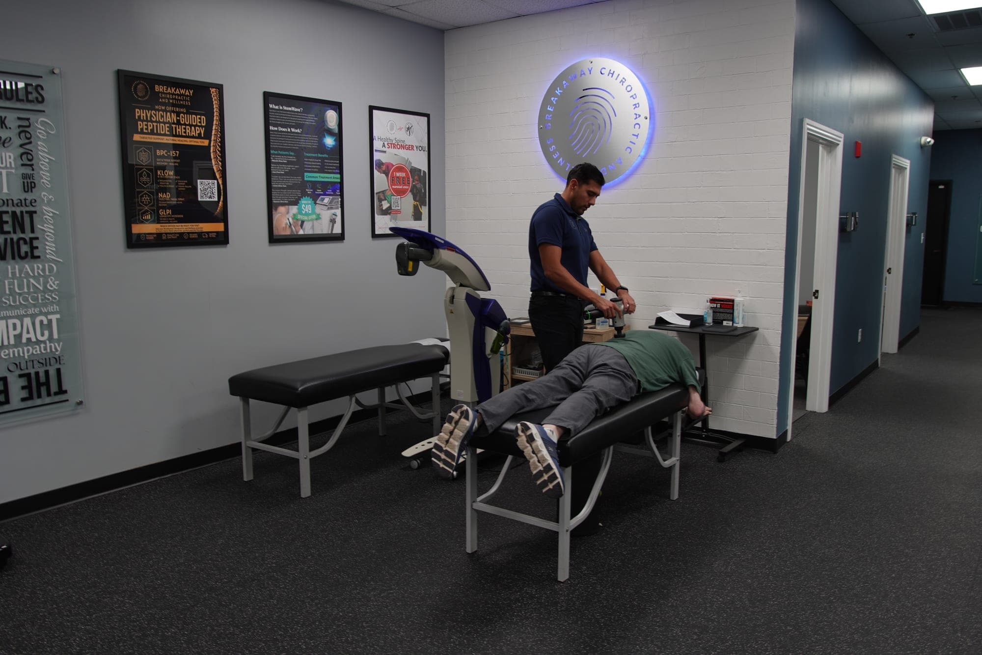 Open treatment area with a patient receiving percussion therapy on a table beneath the Breakaway Chiropractic & Wellness sign